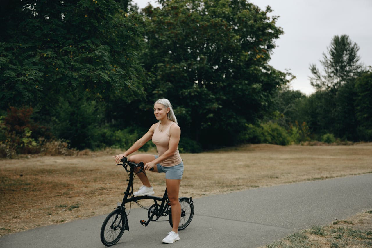 Home A woman in a beige tank top riding a bicycle on a paved path with lush greenery.
